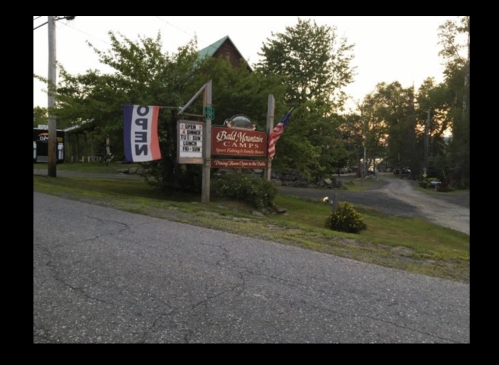 Sign for Bald Mountain Camps with "Open" banner, surrounded by trees and a gravel road at dusk.