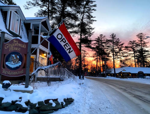 A cozy restaurant with an "OPEN" flag, surrounded by snow and trees, at sunset.