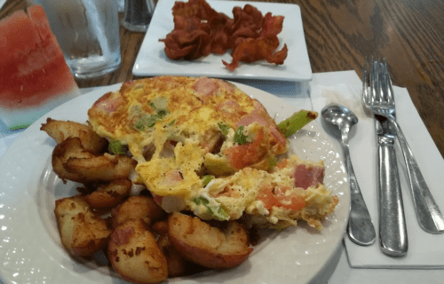 A plate with an omelette, seasoned potatoes, and a side of bacon, accompanied by a slice of watermelon.