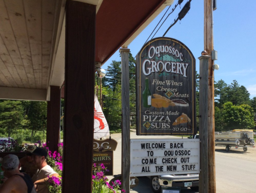 Sign for Oquossoc Grocery featuring fine wines, cheeses, meats, and custom-made pizza and subs, with a welcome message.