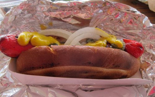 A hot dog in a bun topped with mustard, onions, and relish, wrapped in foil.