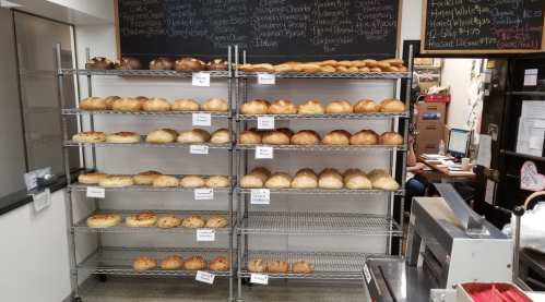 A display of various freshly baked breads on metal shelves in a bakery, with labels for each type.