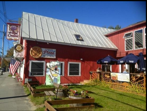 A red building with a metal roof, featuring a pizza sign, outdoor seating, and an American flag. Clear blue sky above.
