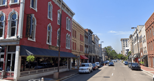 A street view of historic buildings with shops and parked cars under a clear blue sky.