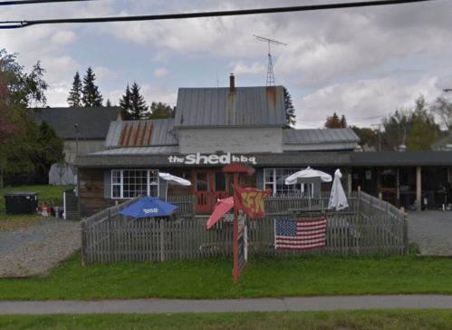 A rustic BBQ restaurant with a wooden exterior, outdoor seating, and a picket fence, featuring umbrellas and an American flag.