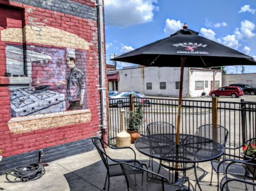Outdoor dining area with a black umbrella, featuring a mural of a chef cooking on a brick wall. Bright blue sky above.
