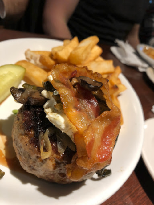 A close-up of a burger topped with bacon and vegetables, served with fries and a pickle on the side.