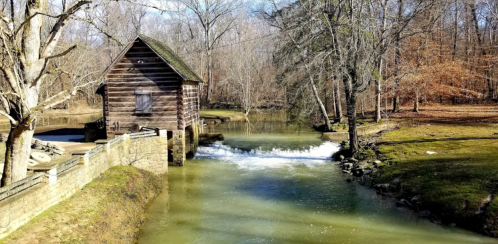 A rustic wooden building by a calm river, with trees and a gentle waterfall in a serene natural setting.
