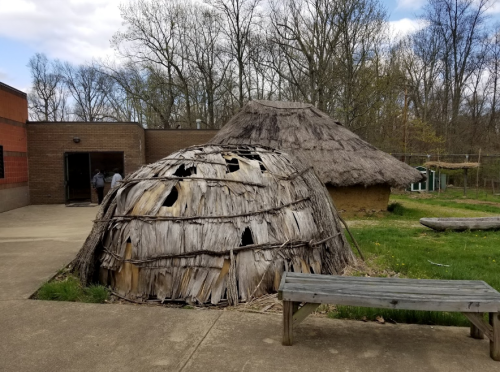 A traditional thatched hut with a rounded shape, surrounded by greenery and a bench, near a building in a wooded area.