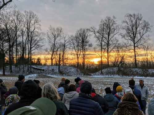 A crowd watches a sunset behind bare trees, with snow covering the ground in a serene winter landscape.