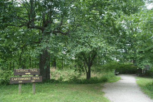 Two wooden signs point to "Terrace Trail" and "Earthworks Trail" amidst lush green trees and a gravel path.