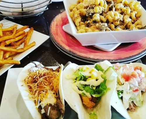 A plate with three tacos filled with various toppings, alongside a bowl of pasta and a side of fries.