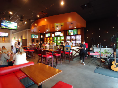 A lively bar interior with red seating, a long counter, and patrons enjoying drinks and food. TVs display sports.
