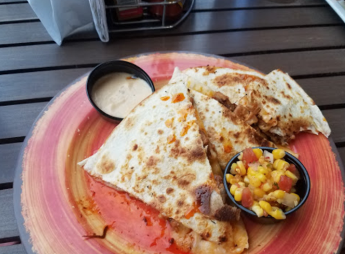 A plate with two quesadilla halves, a small bowl of dipping sauce, and a side of corn salsa on a wooden table.