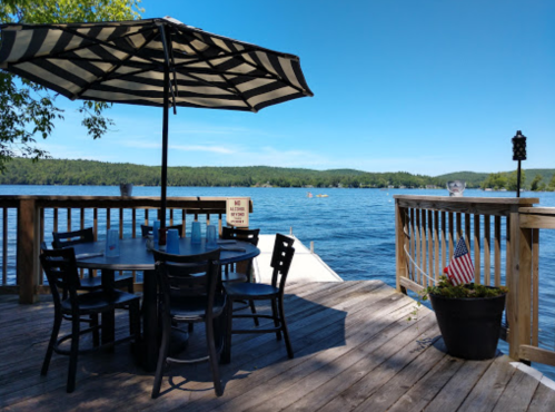 A lakeside deck with a table and umbrella, overlooking calm water and green hills under a clear blue sky.
