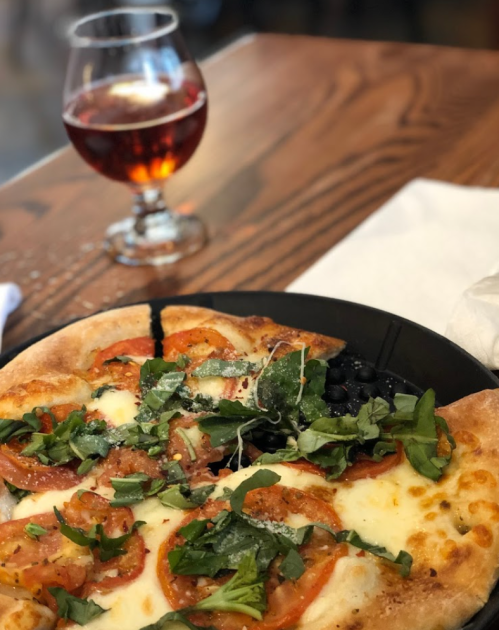 A close-up of a pizza topped with tomatoes, greens, and cheese, alongside a glass of red beverage on a wooden table.