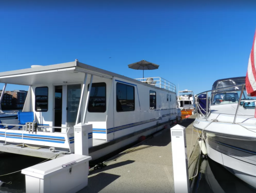 A white houseboat docked at a marina, with a blue sky and other boats nearby.