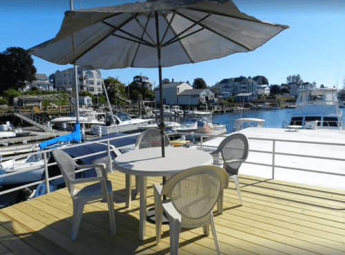 A patio table with chairs and an umbrella overlooks a marina filled with boats and waterfront buildings.