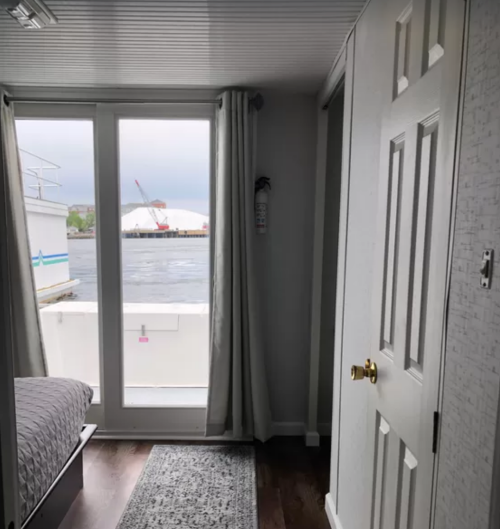 Interior view of a room with a bed, door, and large windows overlooking a body of water and a distant industrial area.