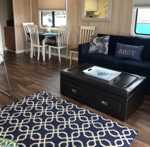 Cozy living space with a blue sofa, coffee table, dining area, and patterned rug. Bright and inviting atmosphere.