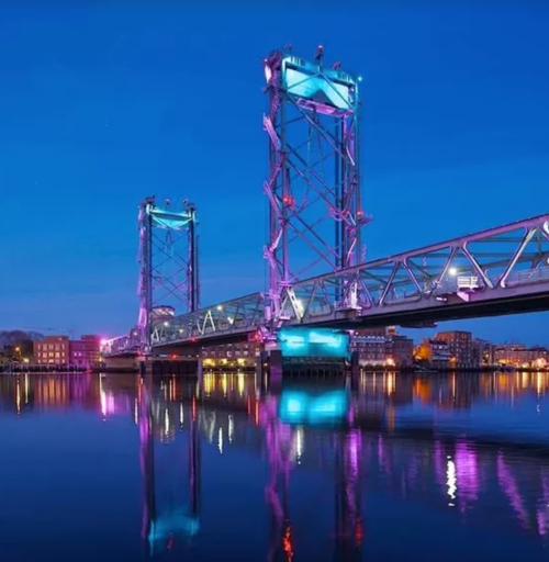 A brightly lit bridge at night, reflecting in calm water, with colorful lights illuminating its structure.