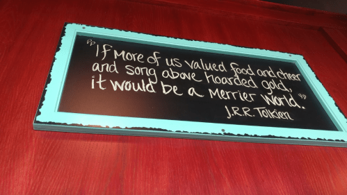 A framed quote on a red wall reads: "If more of us valued food and cheer... it would be a merrier world." - J.R.R. Tolkien.