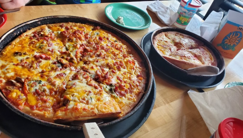Two pizzas on a wooden table: one large with various toppings and a smaller cheese pizza, alongside drinks and plates.