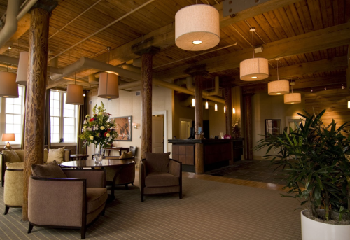 Cozy lobby with wooden beams, plush seating, decorative plants, and warm lighting. A reception desk is in the background.