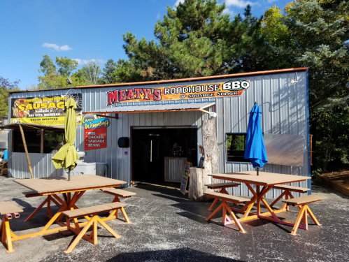 Exterior of Manny's Roadhouse BBQ, featuring picnic tables and colorful umbrellas outside a metal building.