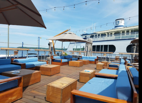 Outdoor seating area with blue cushions and wooden tables, overlooking a calm water view and a boat in the background.