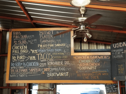 Menu board featuring BBQ items like ribs, brisket, chicken, tacos, and sandwiches, with prices listed.