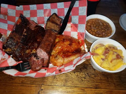 A plate of barbecue featuring ribs, sausage, chicken, baked beans, and peach cobbler, served in a red-checkered basket.