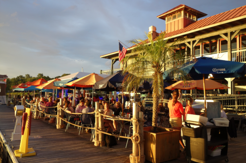 Outdoor dining area with colorful umbrellas, people enjoying meals, and a waterfront view at sunset.