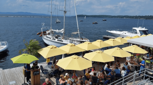 A waterfront scene with boats, yellow umbrellas over a crowded deck, and a sailboat in the background under a blue sky.
