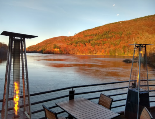 A serene river view at sunset, framed by autumn foliage and outdoor heaters on a deck.