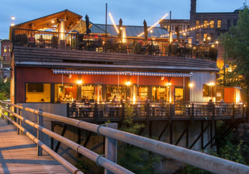 A cozy restaurant with outdoor seating, illuminated by string lights, overlooking a river at dusk.