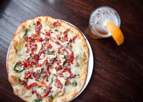 A delicious pizza topped with sun-dried tomatoes, chicken, and cheese, alongside a glass of beer with an orange slice.