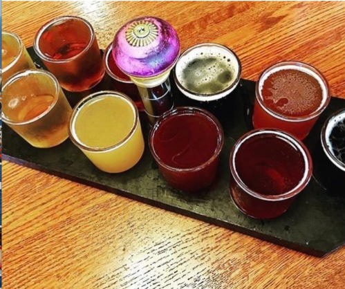 A wooden tray holds ten small glasses of various colorful drinks, including beer and cocktails.