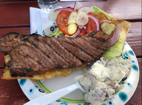 A grilled steak on a bun with lettuce, tomato, onion, and a side of potato salad on a decorative plate.