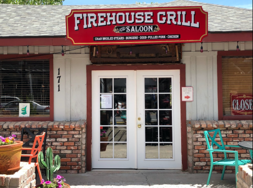 Exterior of Firehouse Grill & Saloon with a red sign, double doors, and colorful chairs outside. Cactus and flowers in view.