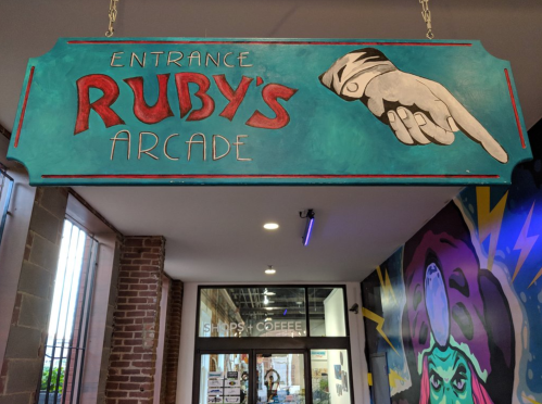 Sign for "Ruby's Arcade" with a painted hand pointing, hanging above an entrance in a colorful corridor.