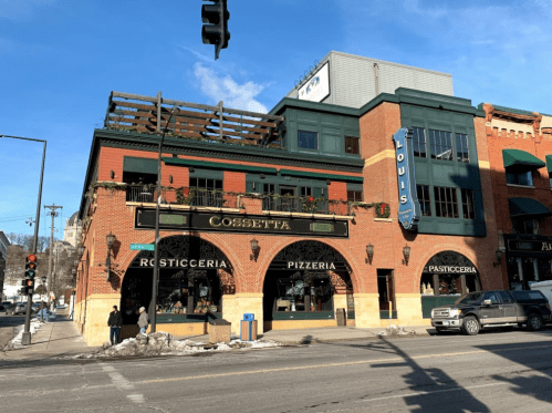 A brick building with a green awning, featuring "Cossetta Rosticceria Pizzeria Pasticceria" signage and outdoor seating.