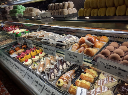 A display case filled with colorful pastries, including macarons, cupcakes, cannoli, and chocolate mousse desserts.