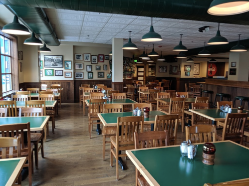 A spacious restaurant interior with wooden tables and chairs, green tabletops, and framed photos on the walls.