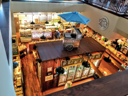 A vibrant market interior with food displays, a cart, and customers browsing under warm lighting.