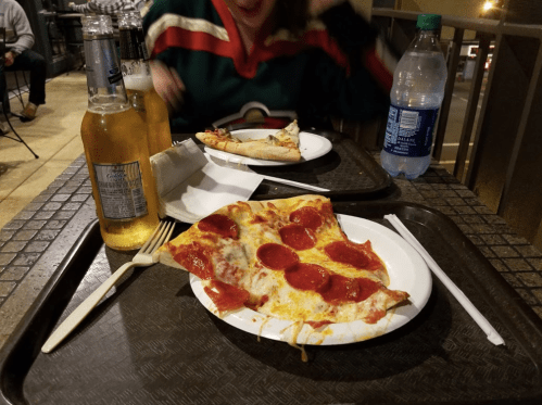 A tray with a slice of pepperoni pizza, a bottle of beer, and a water bottle, with a blurred person in the background.