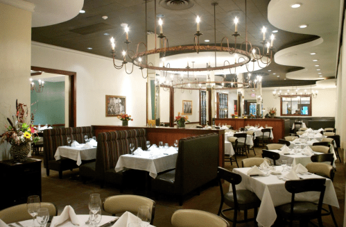 Elegant restaurant interior with white tablecloths, chandeliers, and a cozy seating area. Flowers add a touch of color.