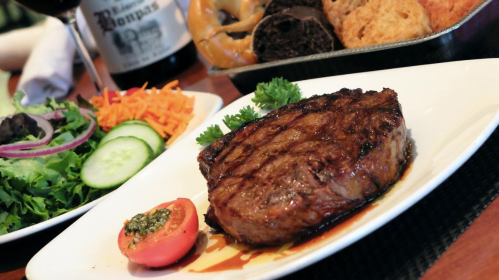 A beautifully grilled steak served with fresh vegetables and sides, including salad and bread, on a white plate.