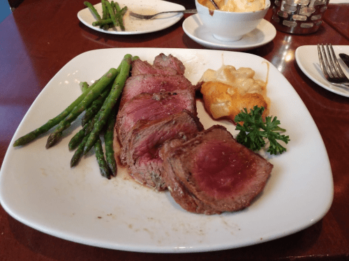 A plate featuring sliced beef tenderloin, asparagus, and a cheesy potato dish, garnished with parsley.