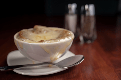 A bowl of creamy soup with melted cheese bubbling over the edge, set on a white plate with a spoon beside it.